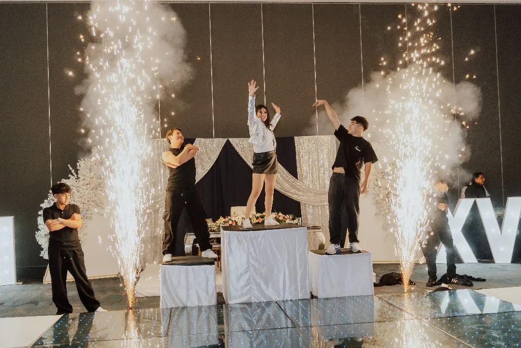 Quinceañera bailando con sus chambelanes sobre tarimas entre fuentes de chispas en su fiesta de XV años.